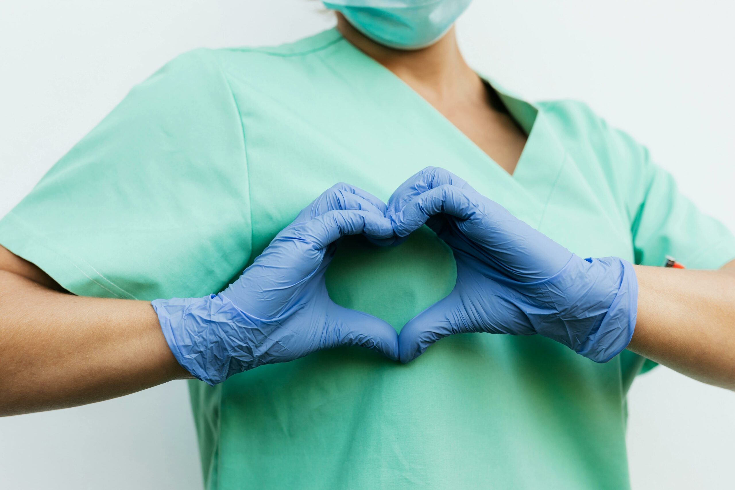 A woman in scrubs and blue gloves forms a heart shape, symbolizing care and compassion in nursing leadership.