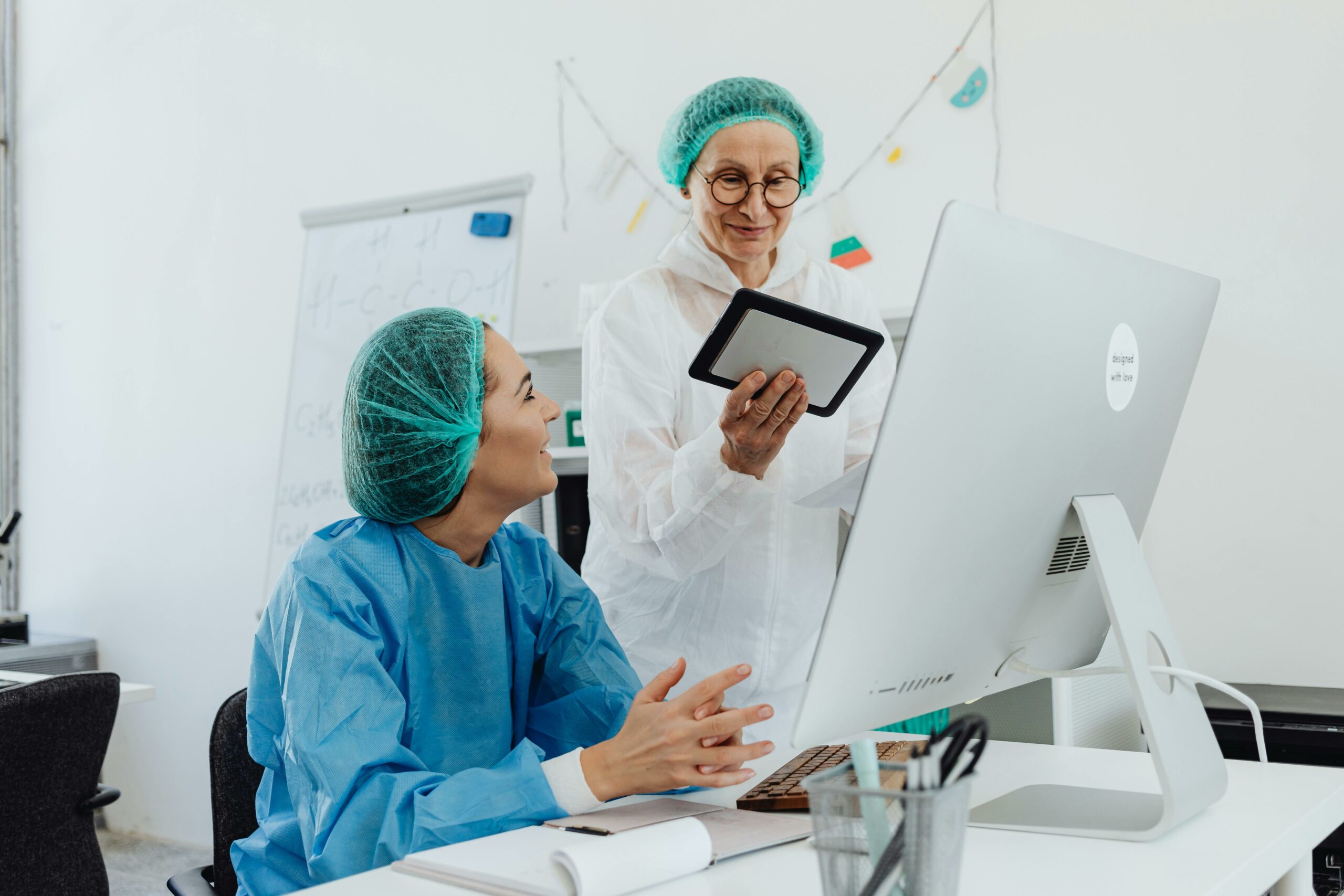 Two women in scrubs collaborate on a computer, focused on their medical transcription service business tasks.