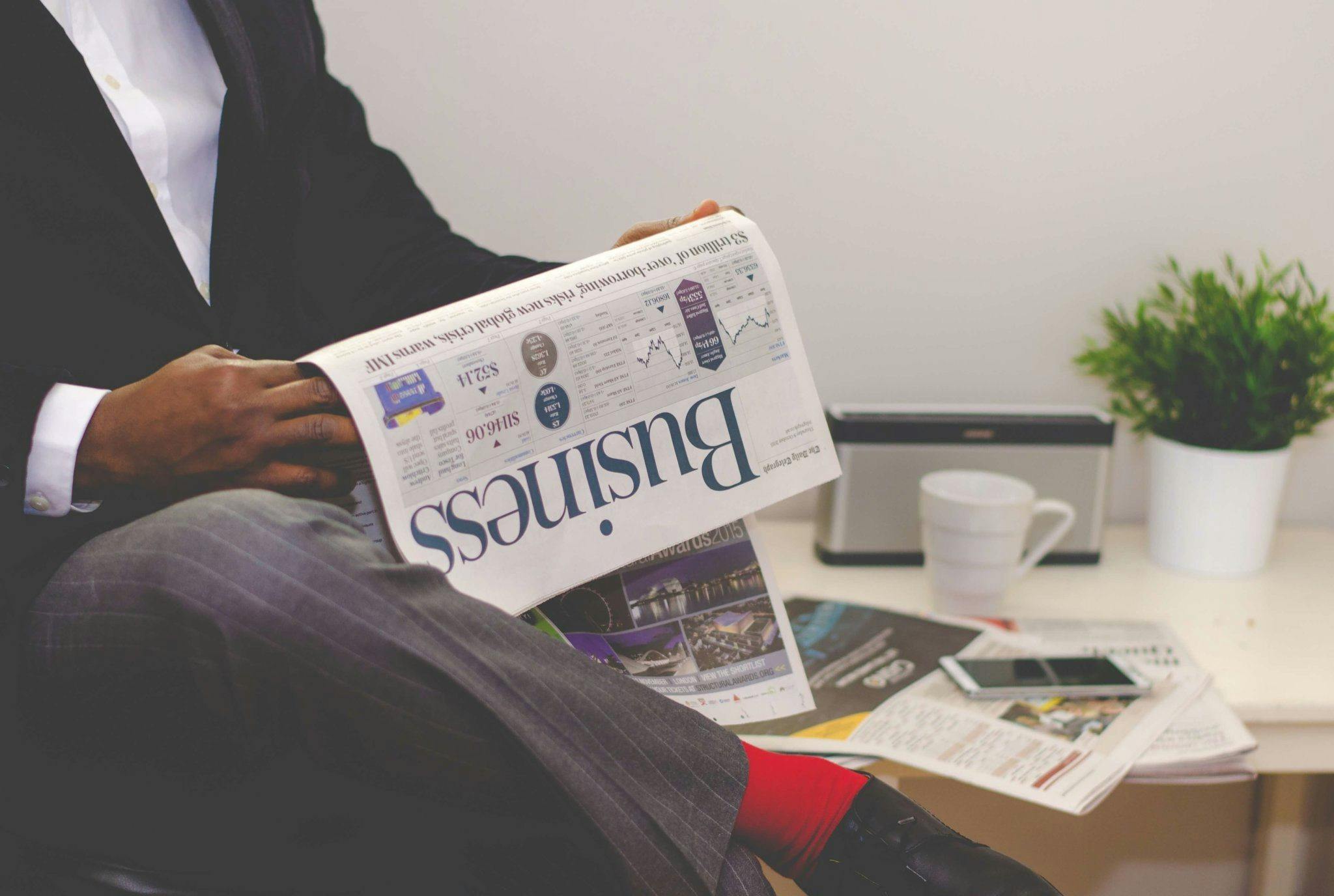 A professional in a suit reading a business newspaper, sitting in an office setting with a plant, a coffee cup, and documents on a table, representing a financial advisor staying informed on market trends.