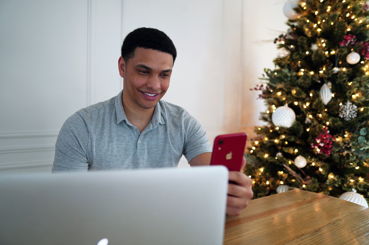 A man, a brokerage services business owner, smiles while using his phone in front of a beautifully decorated Christmas tree.