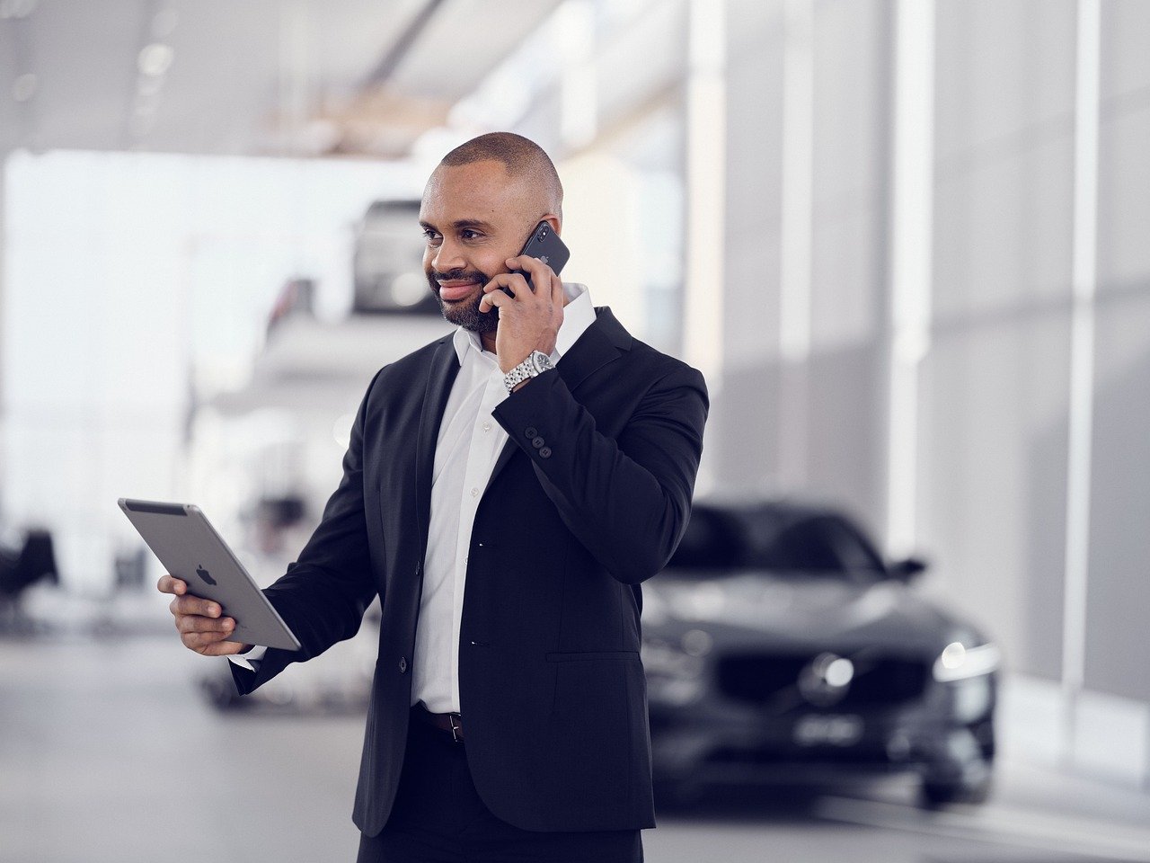 A suited man engages in a phone conversation while standing in-front of a car holding a tablet, symbolizing effective management in automotive services.