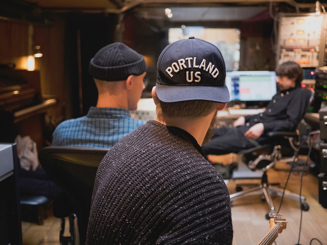 Three men in a recording studio, one wearing a hat, collaborating on a project for their performing arts business.