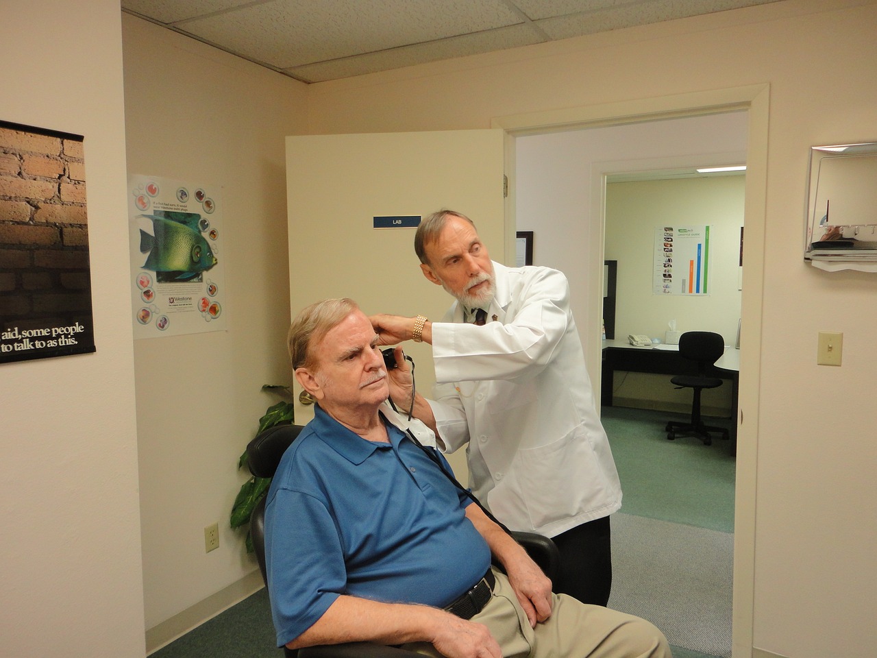 A doctor in a white coat examining an older man's ear with a medical instrument in an audiology clinic room.